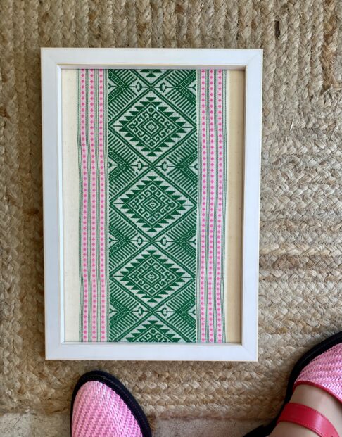 A pea green and baby pink framed textile wall art shown on rush matting.