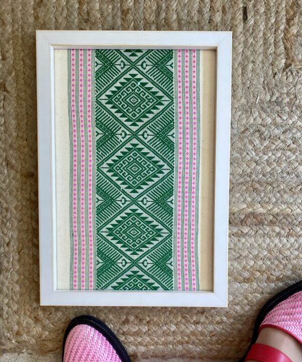 A pea green and baby pink framed textile wall art shown on rush matting.