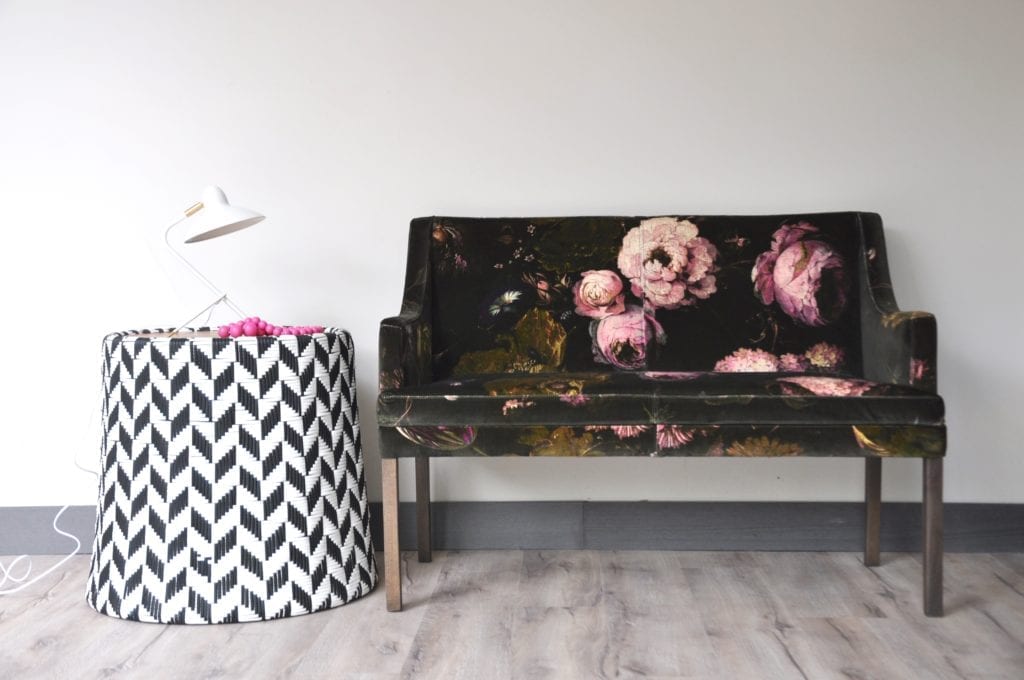 A velvet-upholstered floral bench next to a woven black and white drum side table with modern white metal lamp.