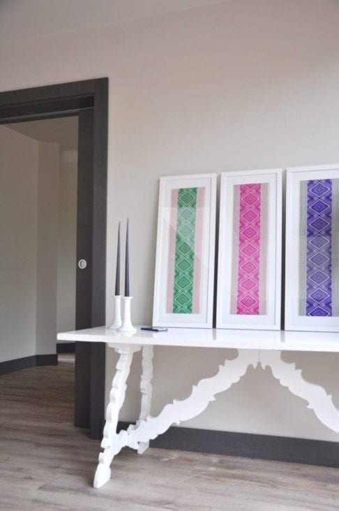 Ready-framed textiles from Thailand in pretty, jewel-box hues, displayed on a white refectory side table in a modern hallway.