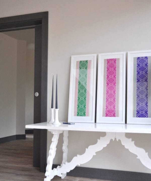 Ready-framed textiles from Thailand in pretty, jewel-box hues, displayed on a white refectory side table in a modern hallway.