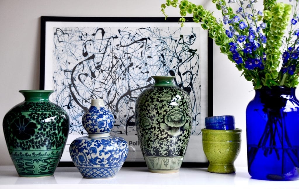 A selection of hand-decorated Thai pottery pots displayed alongside a vibrant floral display in a blue glass vase with a Jackson Pollack print behind.