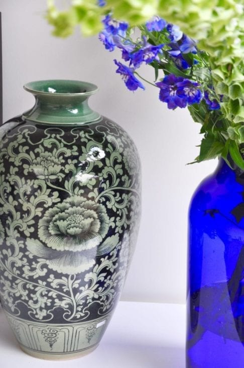 A hand-painted, glazed celadon pot depicting a Lotus flower from northern Thailand. A blue glass vase filled with Delphiniums and Bells of Ireland looks elegant next to it.