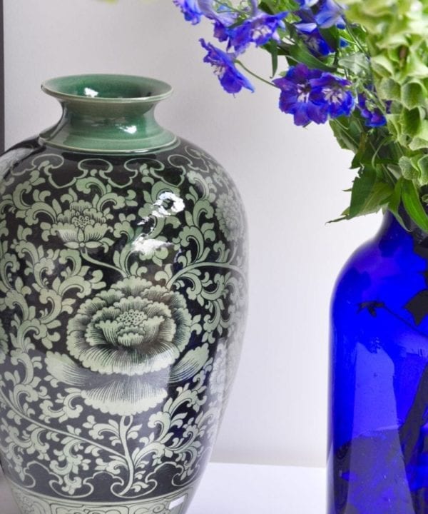 A hand-painted, glazed celadon pot depicting a Lotus flower from northern Thailand. A blue glass vase filled with Delphiniums and Bells of Ireland looks elegant next to it.