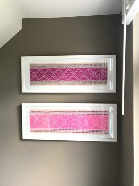 A pair of candy and magenta pink white wood framed textiles hung on a taupe wall by a window.