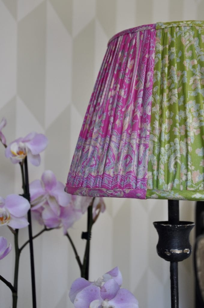 A pink and lime silk gathered lampshade on a simple metal base with modern geometric wallpaper in the background and potted pink orchids.