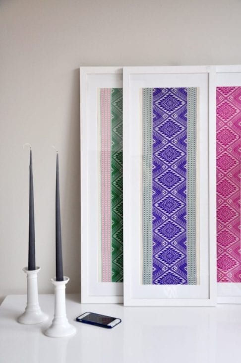 Vibrantly-hued framed textile art, showcasing purple, pink and green Thai cotton wovens framed in tall, white-washed wooden box frames styled on a white table with a pair of candlesticks.