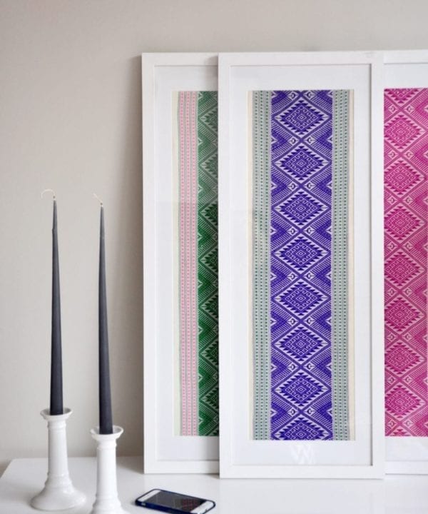 Vibrantly-hued framed textile art, showcasing purple, pink and green Thai cotton wovens framed in tall, white-washed wooden box frames styled on a white table with a pair of candlesticks.