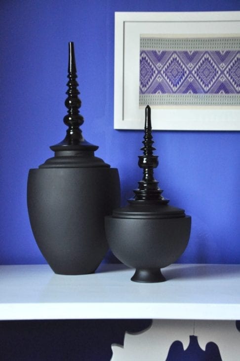 Turned, wooden, lidded pots in matt and gloss black are displayed on a white refectory side table against a Royal blue wall with framed textiles in the background.