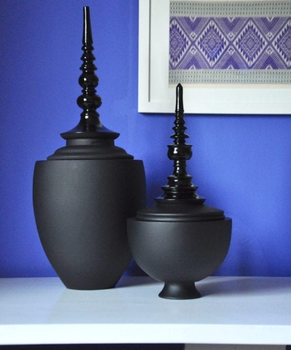 Turned, wooden, lidded pots in matt and gloss black are displayed on a white refectory side table against a Royal blue wall with framed textiles in the background.