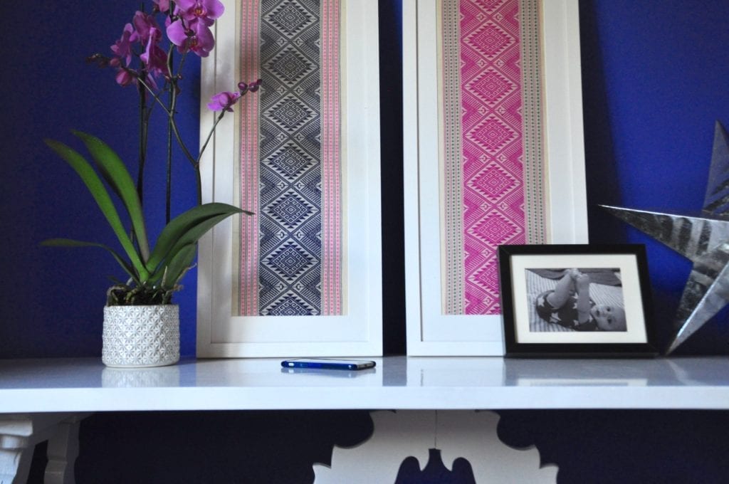 Navy and hot pink Thai textiles framed in white are paired on a sideboard refectory table alongside picture frames, a potted orchid and other decorative objects.