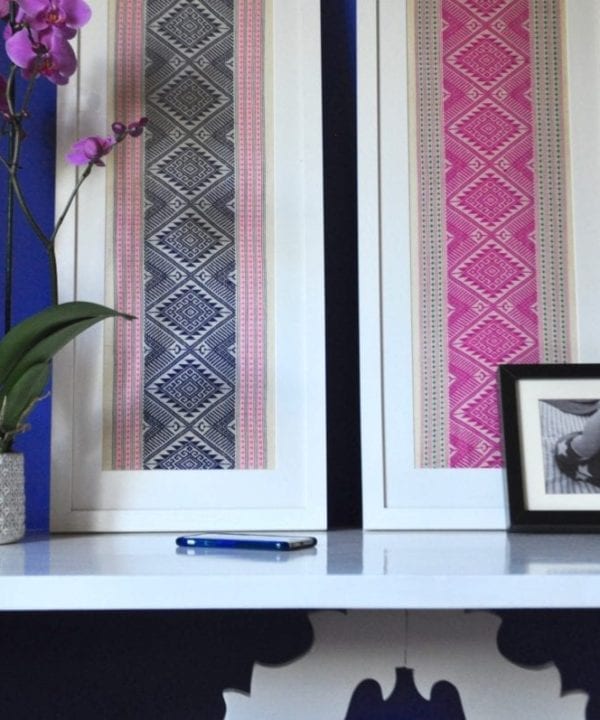 Navy and hot pink Thai textiles framed in white are paired on a sideboard refectory table alongside picture frames, a potted orchid and other decorative objects.