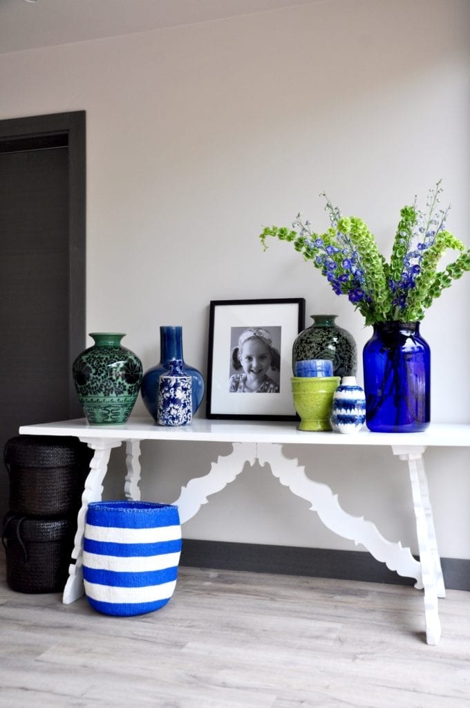 Hand-decorated, oversized pots from Chiangmai in a palette of blues and greens decorate a vintage, refectory style side table in a modern living space. Black and white photography, Delphinium blooms and Bells of Ireland plus a handwoven, striped basket, complete the look.