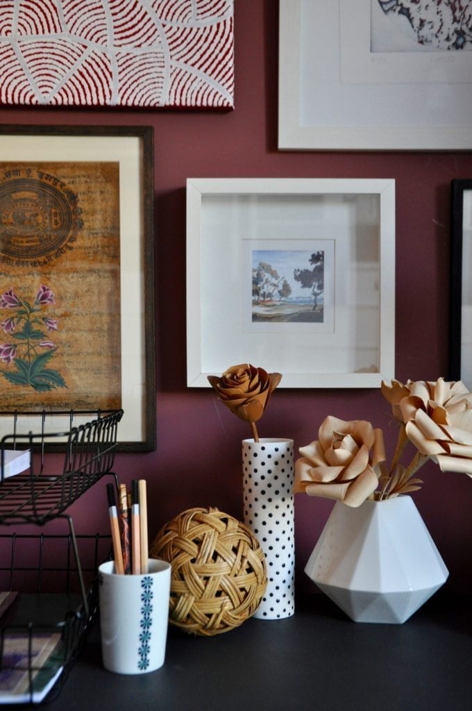 A desktop still life in natural neutrals depicting pen pots, vases, filing trays and travel pictures on an oxblood-red painted wall.