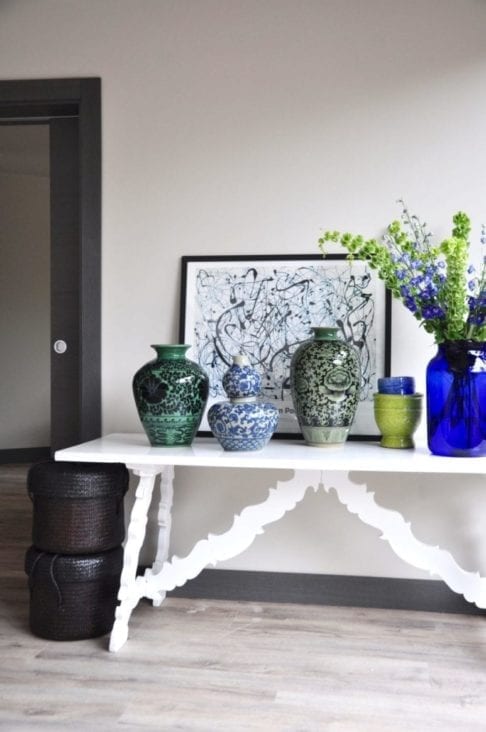 Hand-decorated, oversized pots from Chiangmai in a palette of blues and greens decorate a vintage, refectory style side table in a modern living space. A Jackson Pollack print and a floral display of Delphiniums and Bells of Ireland complete the look.