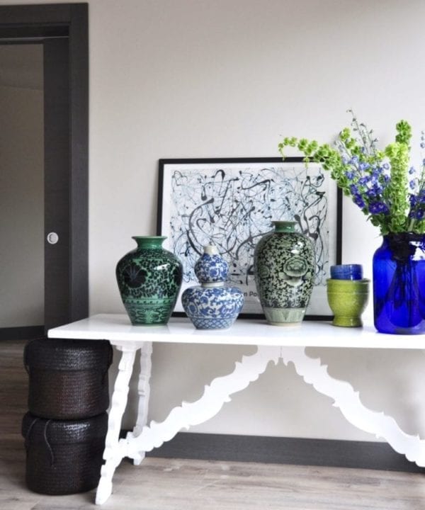 Hand-decorated, oversized pots from Chiangmai in a palette of blues and greens decorate a vintage, refectory style side table in a modern living space. A Jackson Pollack print and a floral display of Delphiniums and Bells of Ireland complete the look.