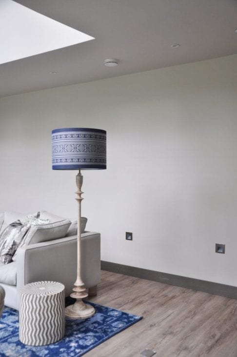 An elegant, Pooky floor lamp is teamed with a large, blue and white drum shade in a Hungarian Folk-inspired stripe design. The setting is a modern sitting room with a decorative blue rug in the foreground.