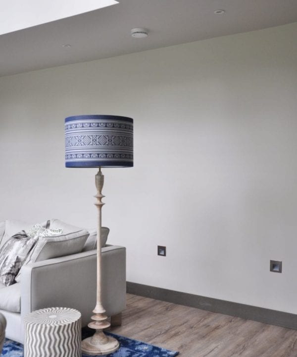 An elegant, Pooky floor lamp is teamed with a large, blue and white drum shade in a Hungarian Folk-inspired stripe design. The setting is a modern sitting room with a decorative blue rug in the foreground.