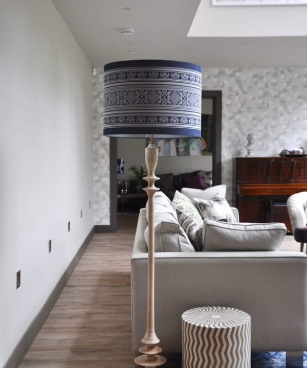 A tall white-washed turned wood floor lamp with patterned blue drum shade at the end of a boxy, modern sofa in a modern, double-height space.