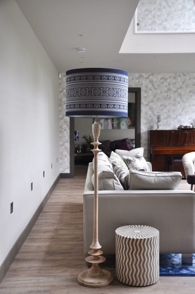 A tall white-washed turned wood floor lamp with patterned blue drum shade at the end of a boxy, modern sofa in a modern, double-height space.