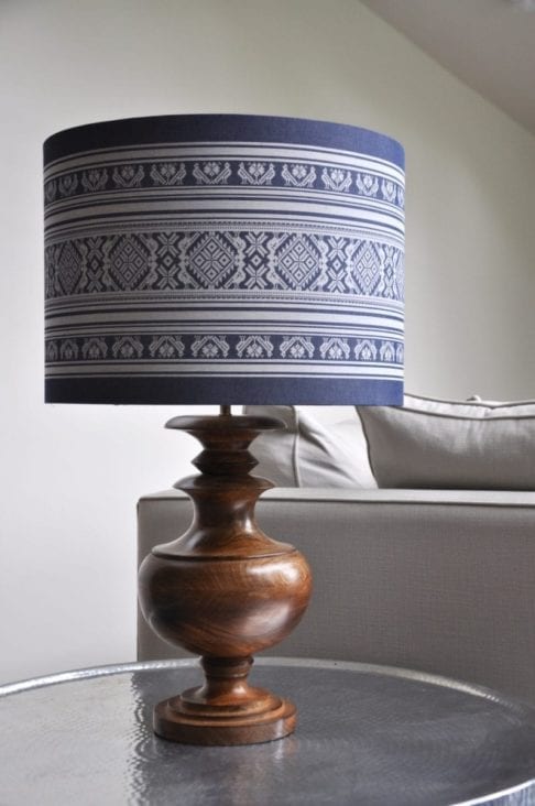 A large blue and white drum lampshade depicting a Hungarian-inspired folk design teamed with a wooden urn lamp base and sat on a beaten metal side table.