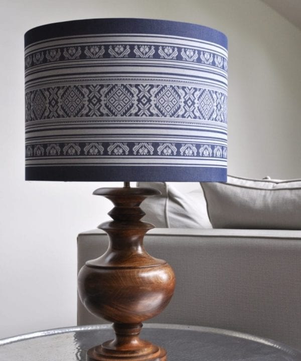 A large blue and white drum lampshade depicting a Hungarian-inspired folk design teamed with a wooden urn lamp base and sat on a beaten metal side table.