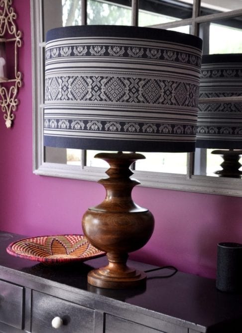 An oversized, blue and white lampshade with a Folk design inspired by traditional Hungarian dress on a wooden urn lamp by Pooky on a sideboard and against a pink-painted backdrop.