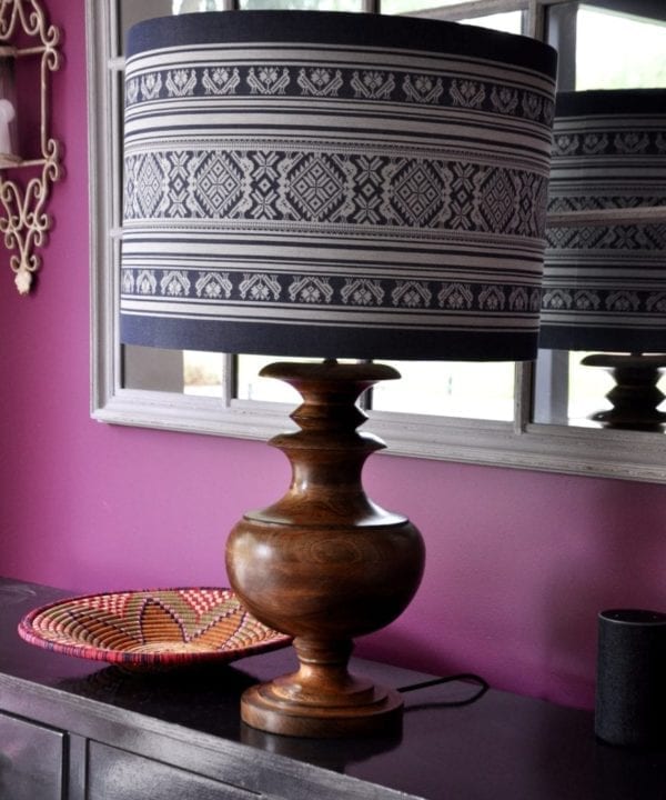 An oversized, blue and white lampshade with a Folk design inspired by traditional Hungarian dress on a wooden urn lamp by Pooky on a sideboard and against a pink-painted backdrop.