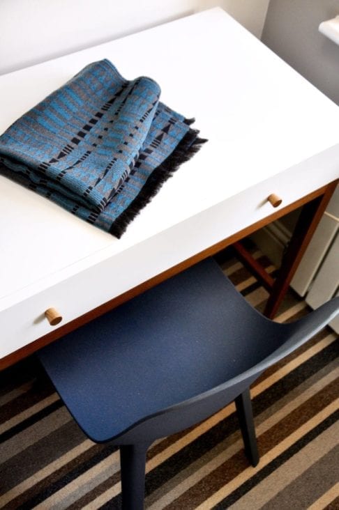 A luxury, cashmere mix, blue and grey scarf in an African-inspire weave on a modern white dressing table in a room with a striped carpet.