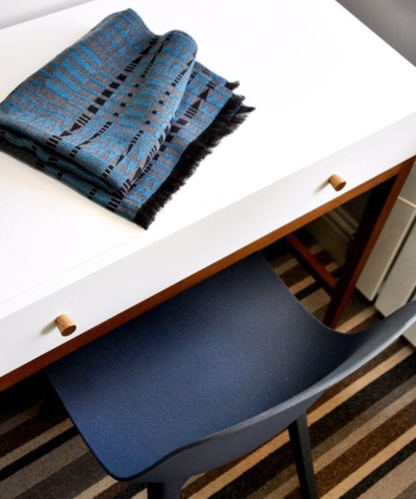 A luxury, cashmere mix, blue and grey scarf in an African-inspire weave on a modern white dressing table in a room with a striped carpet.