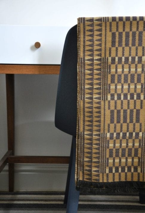 A geometric-weave, cashmere scarf in beiges and browns, hung on the back of a chair in front of a modern, white dressing table.