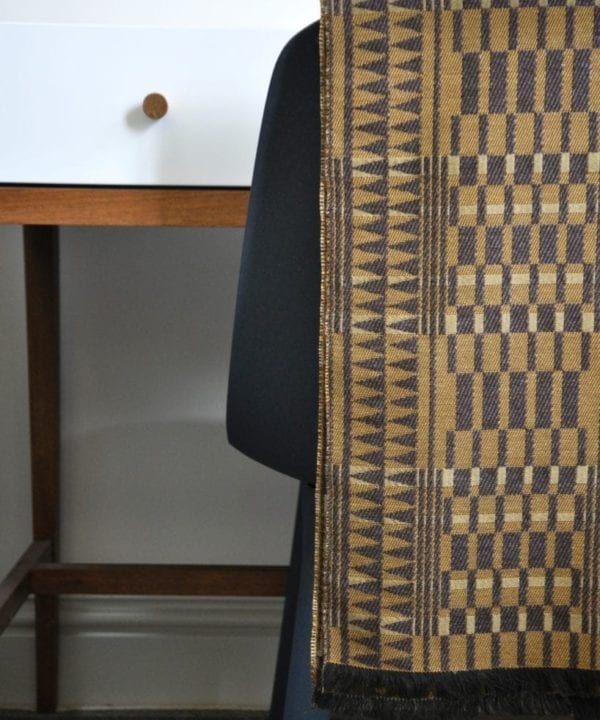 A geometric-weave, cashmere scarf in beiges and browns, hung on the back of a chair in front of a modern, white dressing table.