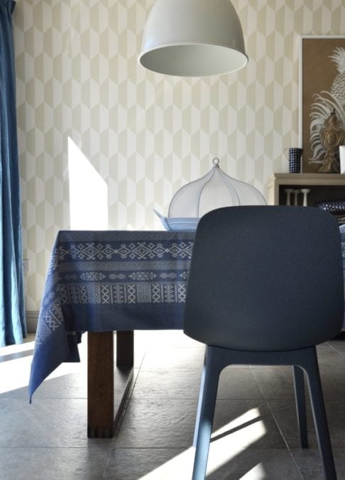 Luxury, patterned, blue linen tablecloth with Arabian motifs on a modern, wooden dining table in a stylish room setting with oversized pendant and geometric wallpaper.