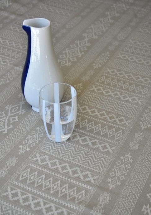 Detail shot of the neutral tablecloth woven with elegant architectural details and shown on a dining table with a white-striped water glass and modern ceramic jug.