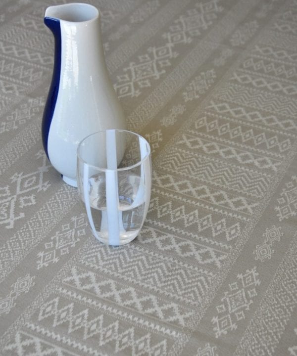 Detail shot of the neutral tablecloth woven with elegant architectural details and shown on a dining table with a white-striped water glass and modern ceramic jug.