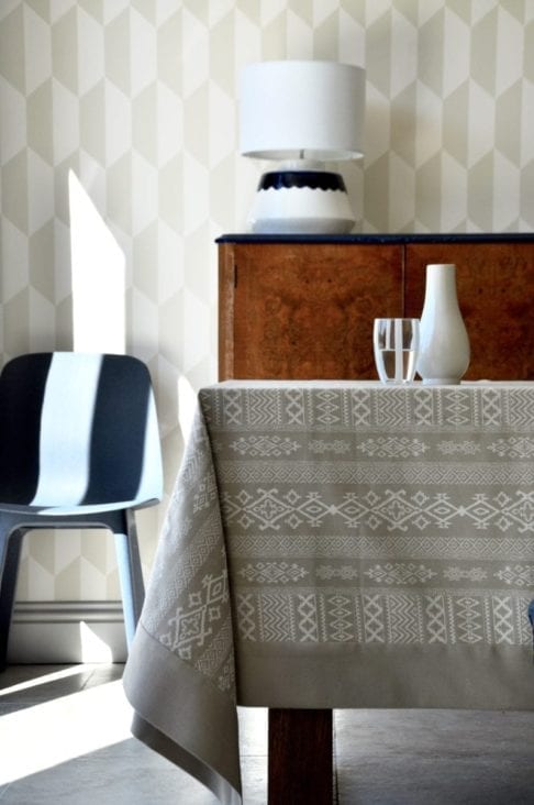 A neutral tablecloth with subtle architectural detailing is styled up in a modern dining room setting with blue chair, brown wood drinks cabinet and geometric wallpaper in the background.