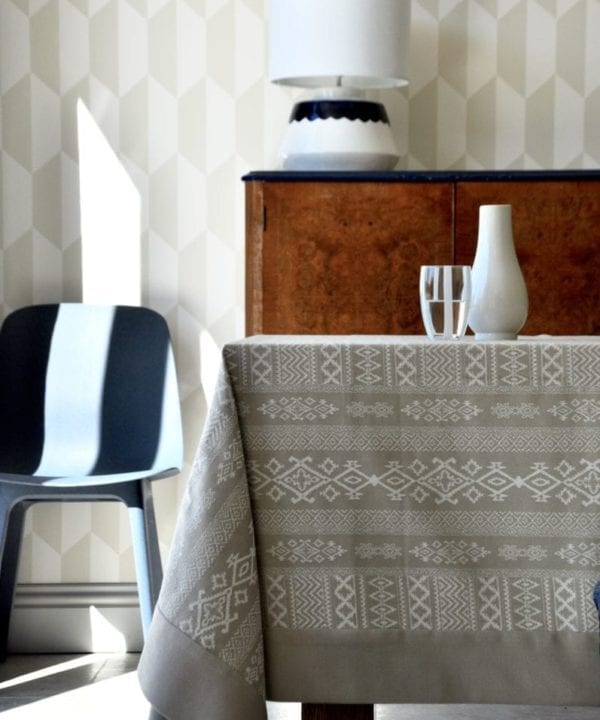 A neutral tablecloth with subtle architectural detailing is styled up in a modern dining room setting with blue chair, brown wood drinks cabinet and geometric wallpaper in the background.