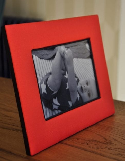 A Thai-silk, red, photograph frame with a picture of a baby on a wooden surface.