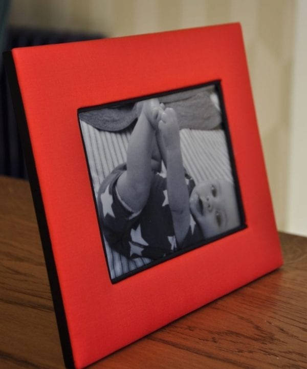 A Thai-silk, red, photograph frame with a picture of a baby on a wooden surface.