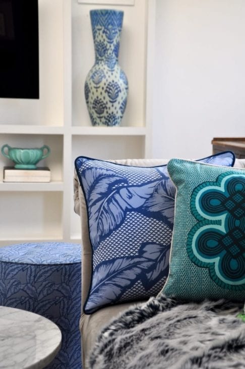 African motif cushion in blue and aqua on a cosy sofa with a faux fur throw and ceramic pots on shelves in the background.