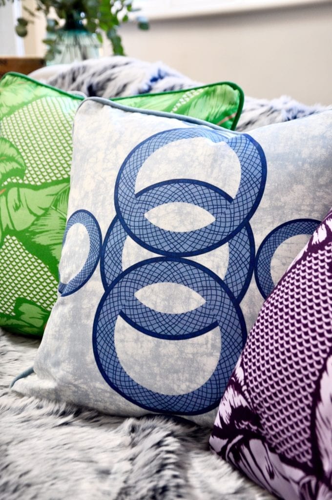 A close up shot of an African circles print cushion in blue with other brightly coloured cushions on a cosy winter sofa.