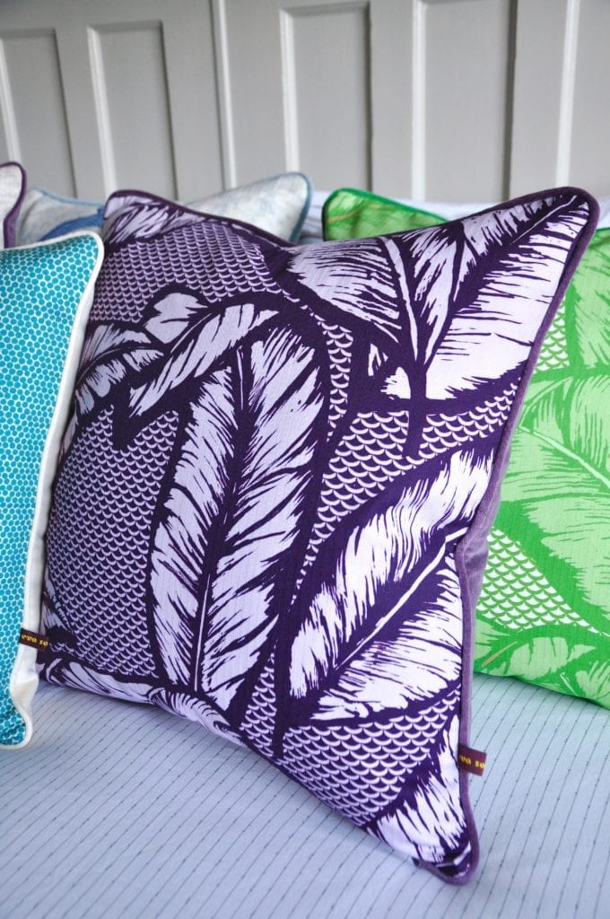 A deep purple Banana leaf print cushion with purple velvet back on a bed with other colourful cushions.