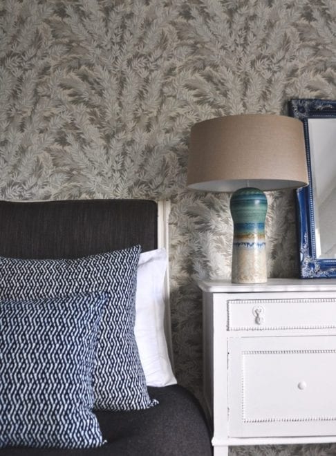 Eastern lattice print cushions in indigo blue dressing a bed, a pottery lamp with linen shade and yew print botanical wallpaper.