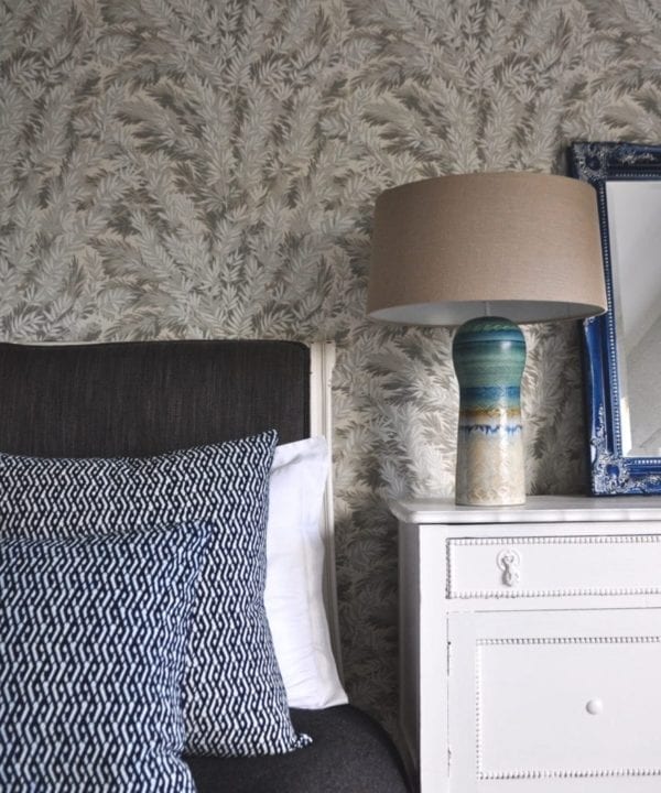 Eastern lattice print cushions in indigo blue dressing a bed, a pottery lamp with linen shade and yew print botanical wallpaper.