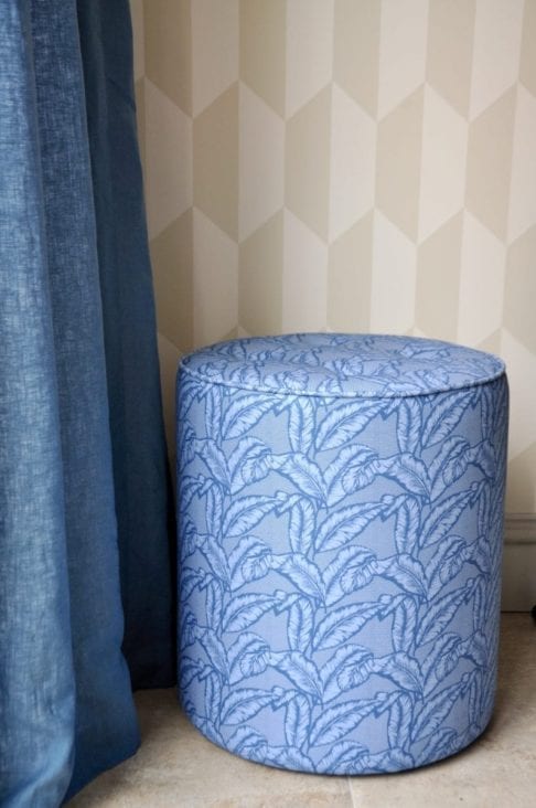 A blue banana print pouffe against neutral geometric wallpaper and next to a linen curtain.