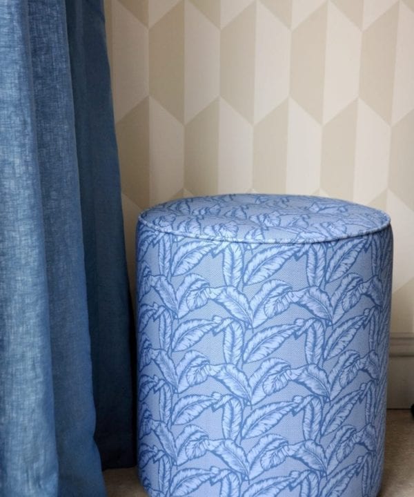 A blue banana print pouffe against neutral geometric wallpaper and next to a linen curtain.