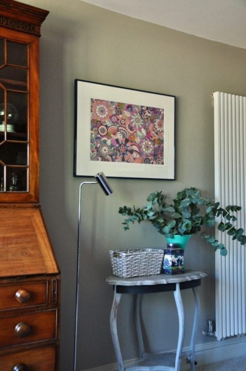 Shot on an angle, a framed, floral, Japanese drawing on a grey wall with a chrome, floor lamp and occasional table with a vase of eucalyptus.