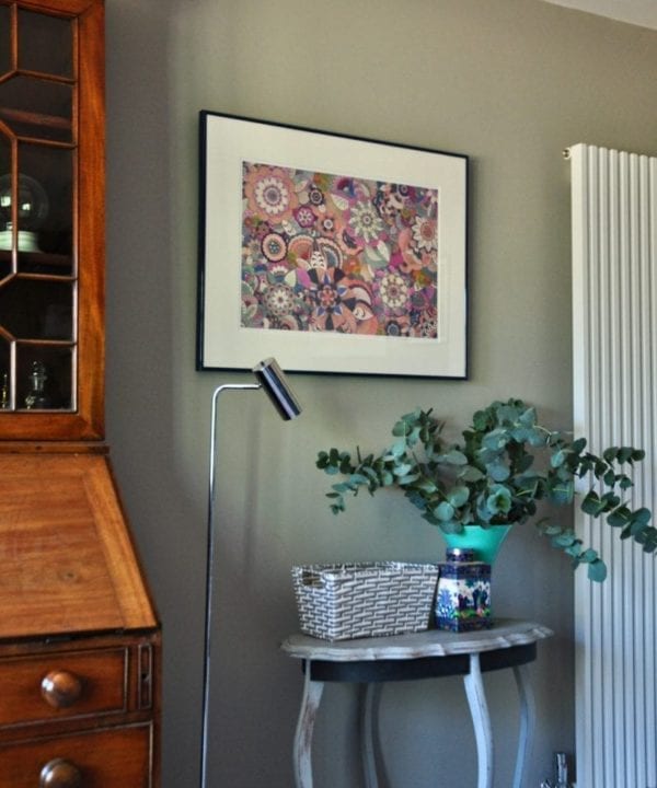 Shot on an angle, a framed, floral, Japanese drawing on a grey wall with a chrome, floor lamp and occasional table with a vase of eucalyptus.