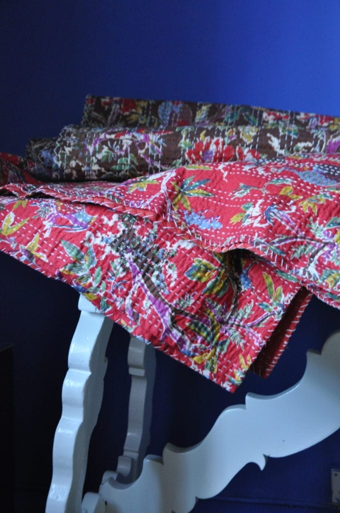 Stitched, red and brown printed bedspreads on a white refectory table against a blue backdrop.