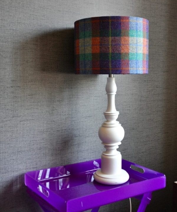 A purple, orange and blue check drum shade made from Harris Tweed on a white wooden lamp base on a purple perspex table. The lampshade is availble to order through Telescope Style.